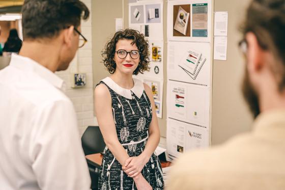 Photo of a female Chatham University student st和ing in front of her presentation, as two professors offer thoughts.